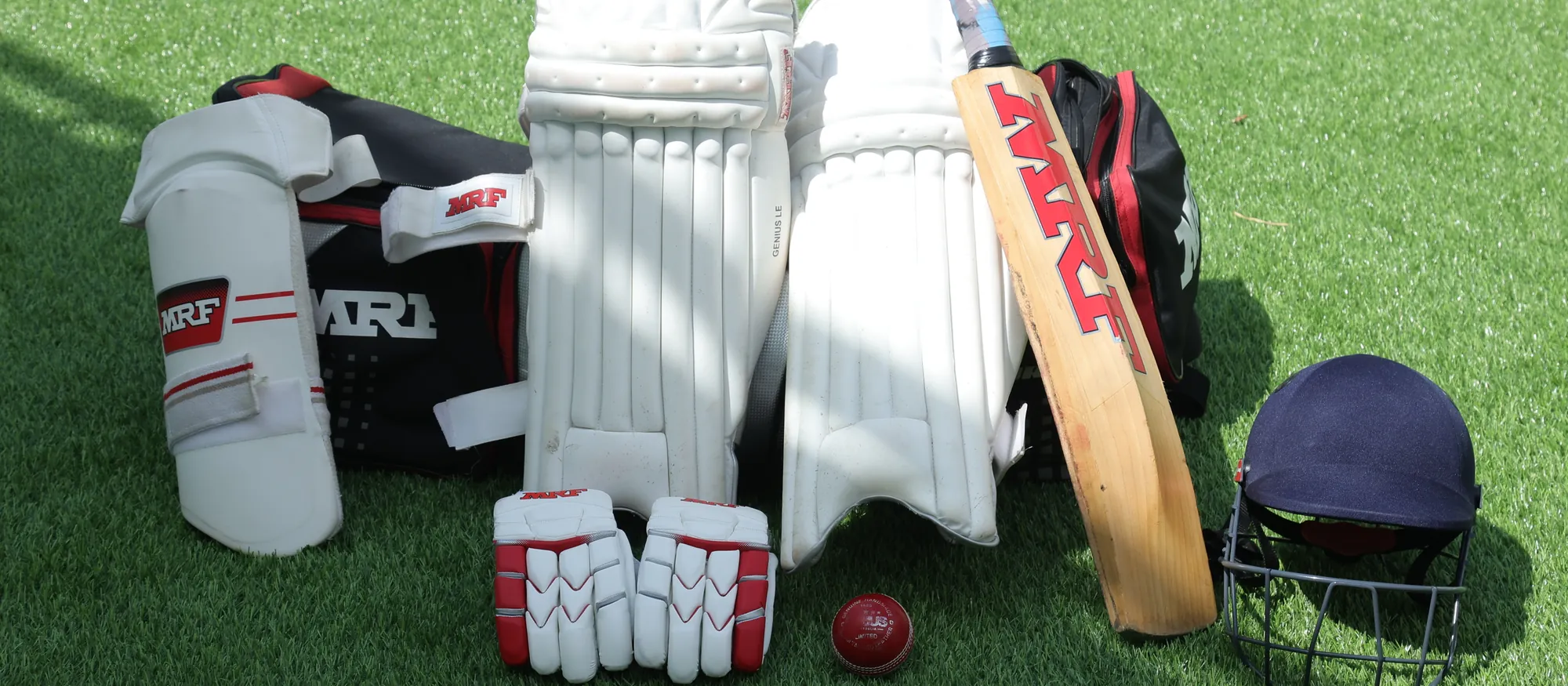 A full set of batting gear laid out on turf: pads, gloves, thigh guard, helmet and an MRF bat.